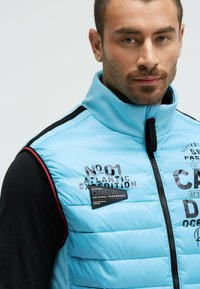 Hellblaues Steppwesten mit schwarzen Akzenten und hohem Kragen. Mit Reißverschluss, Logo-Patches und gestepptem, quilted Design.