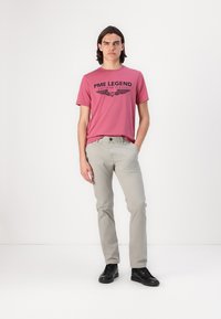 Camiseta de algodón rosa con texto en negro y gráficos de alas, combinada con pantalones gris claro y zapatillas negras. Modelo de pie en un fondo neutro.