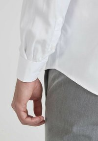 Chemise blanche à manches longues avec poignets boutonnés, texture de tissu lisse, accompagnée de pantalons gris. Le poignet présente un détail de bouton unique.