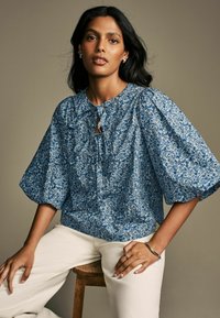 Modra cvetna bluzo z napihnjenimi rokavi, izrezom v obliki ključavnice in nagubanimi detajli. Obuta v kremne hlače, model sedi na leseni klopi.