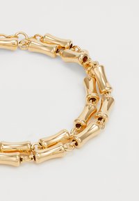 Gouden ketting met verlengde, gladde cilindrische schakels verbonden door kleinere ringen op een lichte achtergrond.