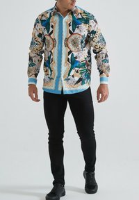 Homme portant une chemise à boutons à motifs avec des horloges, des oiseaux bleus et des motifs floraux, associée à un pantalon slim noir et des chaussures noires.