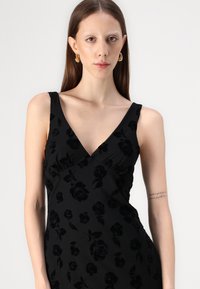 Μαύρο φόρεμα με λουλουδάτη υφή, βαθύ V-ντεκολτέ, εφαρμοστό κορσέ και λεπτές τιράντες. Λείο ύφασμα με διακριτική γυαλάδα.