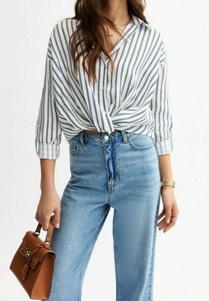 Femme portant une chemise blanche rayée noire nouée devant, jean bleu clair, montre en or, tenant un sac à main en cuir marron à ses côtés.