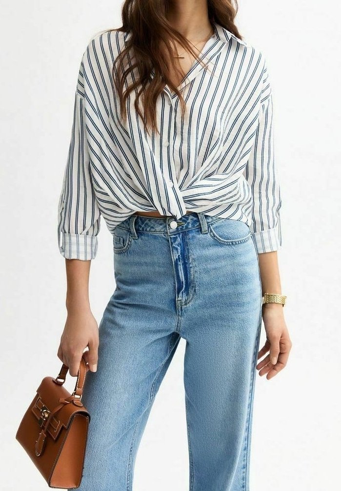 Femme portant une chemise blanche rayée noire nouée devant, jean bleu clair, montre en or, tenant un sac à main en cuir marron à ses côtés.