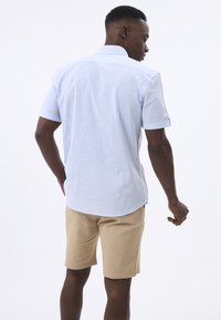 Camisa de manga corta a rayas en azul claro y blanco con un cuello puntiagudo, combinada con pantalones cortos beige. La tela parece suave con un ajuste relajado.