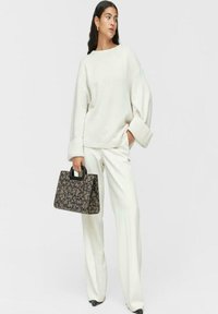 Suéter blanco de gran tamaño con mangas anchas, combinado con pantalones blancos a medida. Lleva un bolso marrón con un patrón y un asa negra.