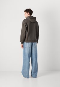 Felpa grigia scura con cappuccio, vestibilità rilassata e orlo a costine, abbinata a jeans a gamba larga blu chiaro e sneakers color crema. Sfondo minimalista.