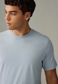 Ung mand med krøllet mørkt hår iført en lyseblå t-shirt med rund hals, der kigger let mod venstre mod en ensfarvet baggrund.