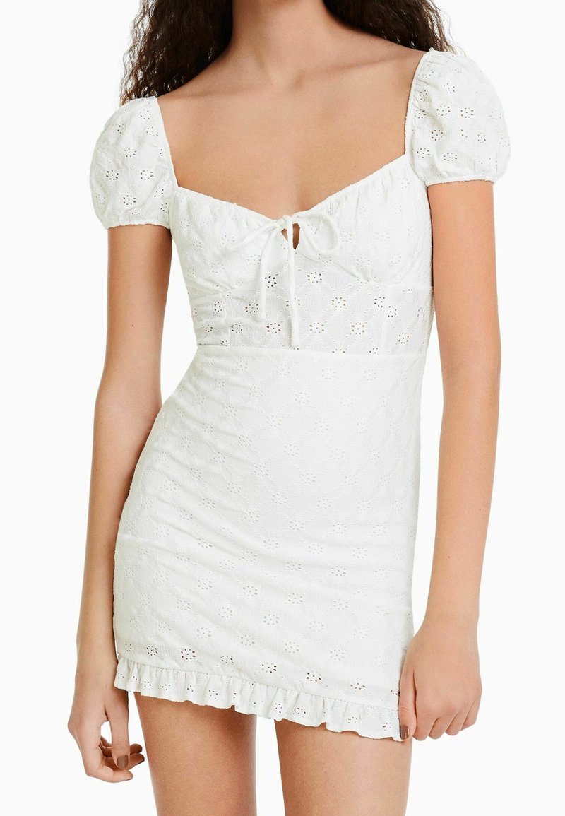 Femme portant une robe courte blanche en broderie anglaise, manches bouffantes, détail noué à l'avant et ourlet à volants.