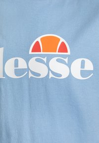 Světle modré tričko s výrazným bílým textem slova "lesse" a nad ním polooblačný oranžový a červený grafický prvek. Měkká textilie.