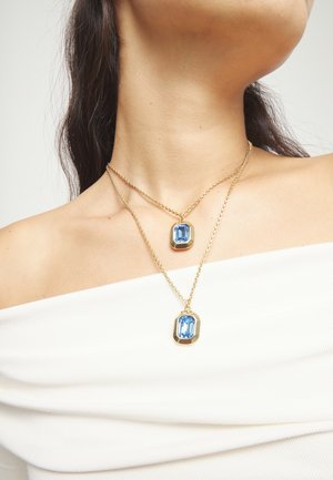 Deux colliers en or avec des pendentifs rectangulaires en pierre précieuse bleue portés au cou d'une femme sur un haut blanc à épaules dénudées.