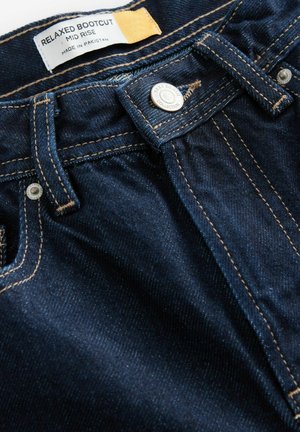 Jeans en denim bleu foncé avec une coupe à taille mi-haute, design bootcut décontracté, couture dorée et fermeture à bouton en métal. L'étiquette indique "Fabriqué au Pakistan."