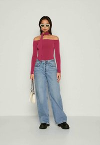 Vrouw in een off-shoulder, lange mouwen, roze top, met wijde blauwe jeans, witte zonnebril, zwarte schoenen en een kleine witte handtas.