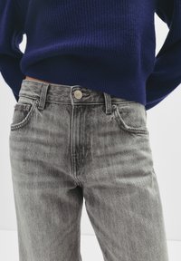 Jean en denim gris avec une finition légèrement délavée, un design à cinq poches, et une fermeture par bouton argenté à la taille, assorti à un pull en maille bleu marine.