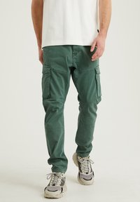 Gröna cargobyxor med en avslappnad passform, som har flera fickor, sömnadsdetaljer och ett mjukt, texturerat tyg. Bärs med grå sneakers.