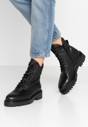 Bottines noires en cuir à lacets avec semelles robustes portées avec un jean bleu clair retroussé sur fond blanc.