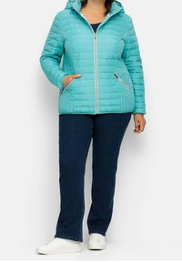 Tyrkysová prošívaná bunda s kapucí, předním zipem a dvěma bočními kapsami. Kombinována s tmavě modrými džíny a bílými teniskami s texturovaným povrchem.