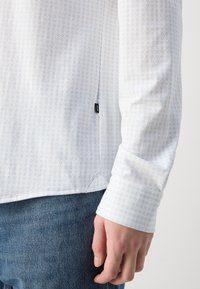 Camisa branca com um padrão geométrico azul claro, mangas longas e punhos com botões. Apresenta uma fenda lateral e uma pequena etiqueta da marca na bainha.