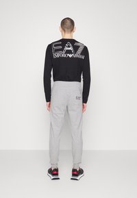 Černé tričko s dlouhým rukávem s bílými logy EA7 na zádech, doplněné šedými tepláky s logem na zadní kapse a černými teniskami.