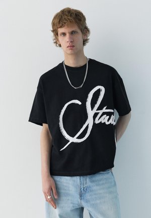 Young man with light brown hair wearing a black oversized T-shirt with bold white script and light blue jeans, standing against a white wall.