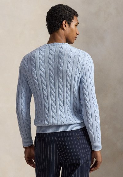 Άνδρας που φοράει γαλάζιο πλεκτό πουλόβερ με σχέδιο σεντονιού και σκουρόχρωμο παντελόνι με ρίγες, από πίσω, μπροστά σε ένα απλό φόντο.