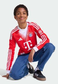 Rode Bayern München-jersey met witte accenten, logo en drie-strepen ontwerp. Gecombineerd met blauwe spijkerbroek en zwarte sneakers met een lichte zool.