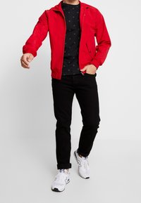 Röd zip-up jacka, svart mönstrad T-shirt, svarta jeans och vita sneakers. Jackan har en ståkrage och framfickor.