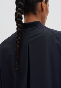 Marineblauwe jas met een hoge kraag, een gladde textuur en een plooi aan de achterkant. Heeft een gevlochten haar detail zichtbaar aan de achterkant.