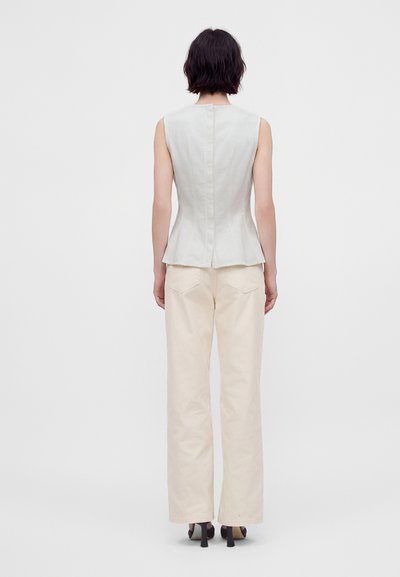 Femme aux cheveux courts et foncés portant un haut blanc moulant sans manches et un pantalon large crème, debout devant un fond clair uni.