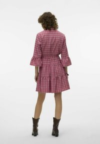 Rozā rakstaina kleita ar sašaurinātu jostu, platiem piedurknēm un slāņotu svārku, izgatavota no vieglas auduma. Valkāta ar brūnām papēžu zābakiem.