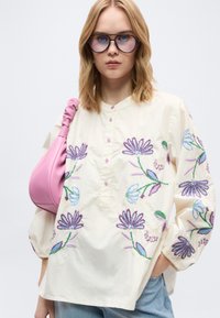 Femme blonde portant de grandes lunettes rondes, un chemisier crème brodé de fleurs violettes et bleues, un jean bleu clair et un sac à main rose.