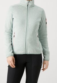 Veste polaire bleu clair à fermeture éclair, avec des détails de fermeture éclair roses, portée avec un pantalon noir ajusté.