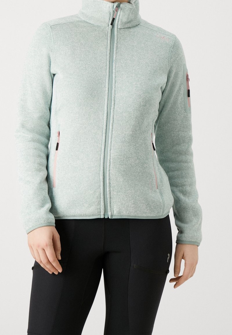 Veste polaire bleu clair à fermeture éclair, avec des détails de fermeture éclair roses, portée avec un pantalon noir ajusté.