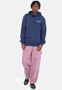 Sudadera azul marino con bolsillo delantero y logotipo en color teal, combinada con pantalones cargo rosa claro con paneles cosidos y bolsillos laterales. Zapatillas negras.