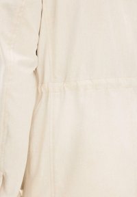Primo piano di tessuto beige con cuciture visibili e una cucitura cucita, che mostra parte della manica e del pannello laterale di un capo d'abbigliamento.