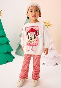 Felpa bianca con una grande grafica di Minnie Mouse con cappello da Babbo Natale, abbinata a pantaloni svasati a quadretti rossi e sneakers bianche. Sullo sfondo alberi verdi e bianchi.