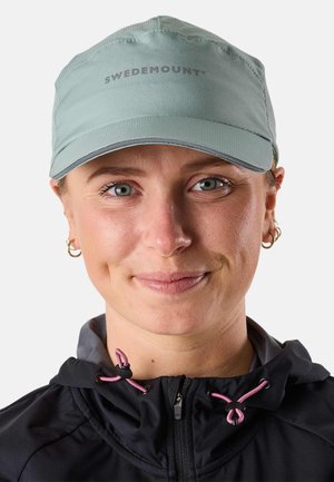 Femme portant une casquette vert clair avec le texte « SWEDEMOUNT », de petites boucles d'oreilles créoles et une veste noire avec des cordons de serrage roses, souriant légèrement.