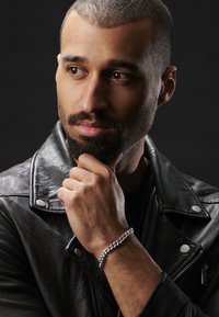 Black leather jacket with silver hardware, black T-shirt underneath, and a silver chain bracelet on the left wrist. Short, groomed hair.