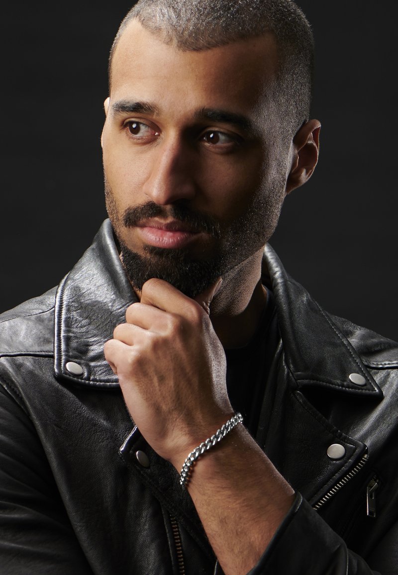 Black leather jacket with silver hardware, black T-shirt underneath, and a silver chain bracelet on the left wrist. Short, groomed hair.