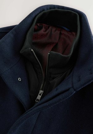 Cappotto in misto lana blu navy con colletto a coste nero, chiusura con zip e rivestimento testurizzato bordeaux, caratterizzato da un dettaglio di design cucito.