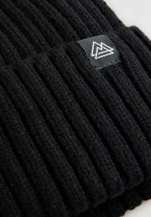 Bonnet en tricot noir avec une texture à côtes verticales, présentant une étiquette tissée affichant un logo géométrique avec des accents blancs et gris.