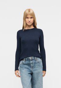 Νεαρή γυναίκα με ίσια ξανθά μαλλιά, που φοράει εφαρμοστή μπλε σκούρη μπλούζα με μακριά μανίκια και άνετο ανοιχτό μπλε τζιν, στέκεται μπροστά σε λευκό φόντο.