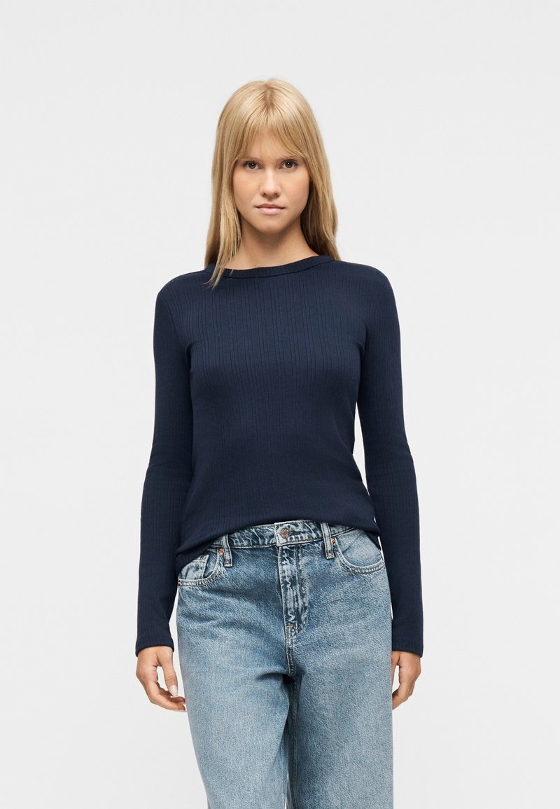 Νεαρή γυναίκα με ίσια ξανθά μαλλιά, που φοράει εφαρμοστή μπλε σκούρη μπλούζα με μακριά μανίκια και άνετο ανοιχτό μπλε τζιν, στέκεται μπροστά σε λευκό φόντο.