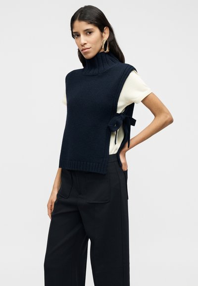 Μπλε ναυτικό πλεκτό πουλόβερ χωρίς μανίκια με ρόλο λαιμό και λεπτομέρεια δεσίματος στο πλάι, συνδυασμένο με κρεμ κοντομάνικο τοπ και μαύρο παντελόνι με ψηλή μέση.