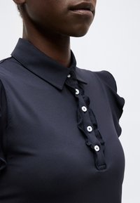 Άτομο που φοράει μπλε σκούρο πουκάμισο με γιακά, με πιέτες στο σκέλος και τέσσερα άσπρα κουμπιά, πάνω σε απλό φωτεινό φόντο.