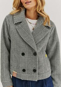 Čierno-biela bunda s rybími kostrami, veľkými klopami, dvojradovým gombíkom a bočnou nášivkou; vyrobená z texturovanej látky.