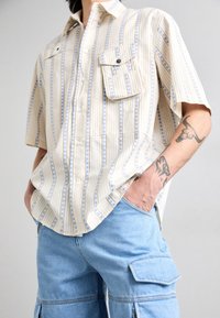 Short-sleeve button-up shirt in beige with vertical blue stripes and small floral patterns. Features two chest pockets and a relaxed fit.
