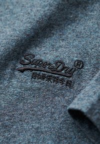 Tessuto textured blu scuro con un logo ricamato "SuperDry" in nero. Aspetto leggermente melange con una texture liscia e morbida.