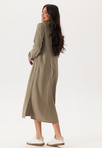Olive green sukienka z długim rękawem, z teksturowanej tkaniny, z bocznym rozcięciem, noszona z białymi sneakersami, ukazująca luźny krój.